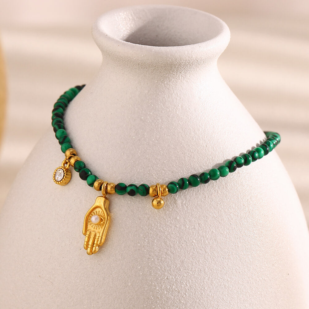 Green beaded bracelet with gold hand charm on a beige vase.